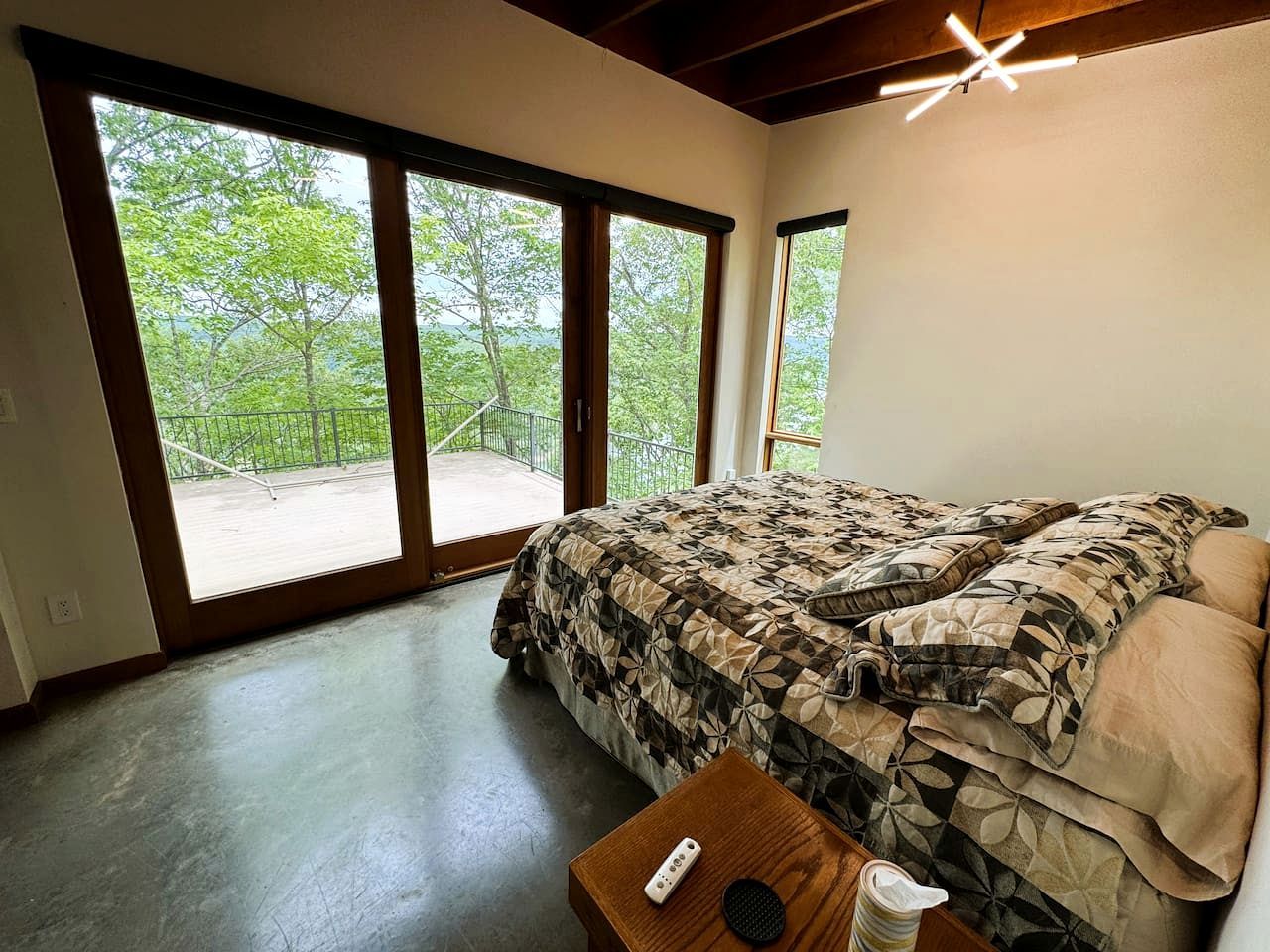 Een slaapkamer met een groot bed, een houten nachtkastje, een afstandsbediening en uitzicht op een balkon met beplanting.