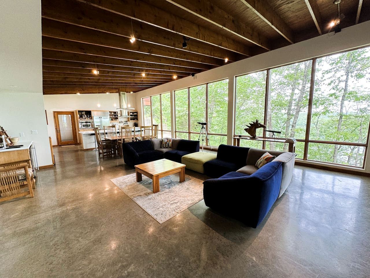 A spacious living area with a large window, a wooden ceiling, and a kitchen in the background.