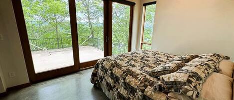 Un dormitorio con una cama grande, una mesita de noche de madera, un control remoto y vistas a un balcón con vegetación.