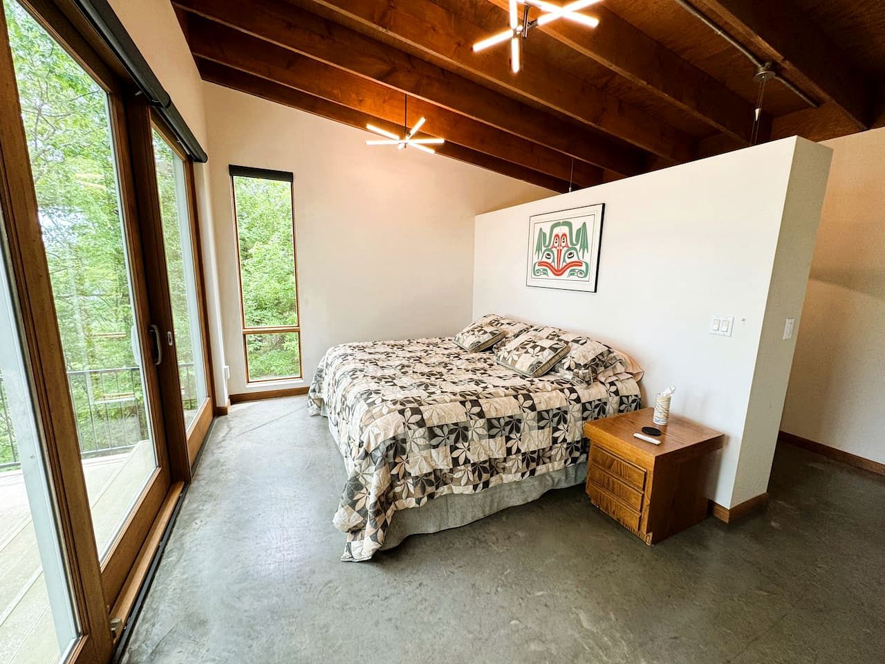 A bedroom with a large bed, a nightstand, a wall-mounted artwork, and a view through a large window.