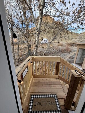Une terrasse en bois avec un paillasson, une vue sur un paysage aride à la végétation clairsemée et un ciel dégagé.