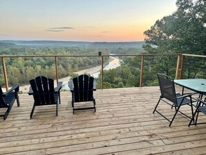 Une terrasse en bois, avec des chaises et une table, donnant sur une rivière et un paysage boisé.