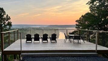 A deck with Adirondack chairs and a table, overlooking a body of water and distant landscape.