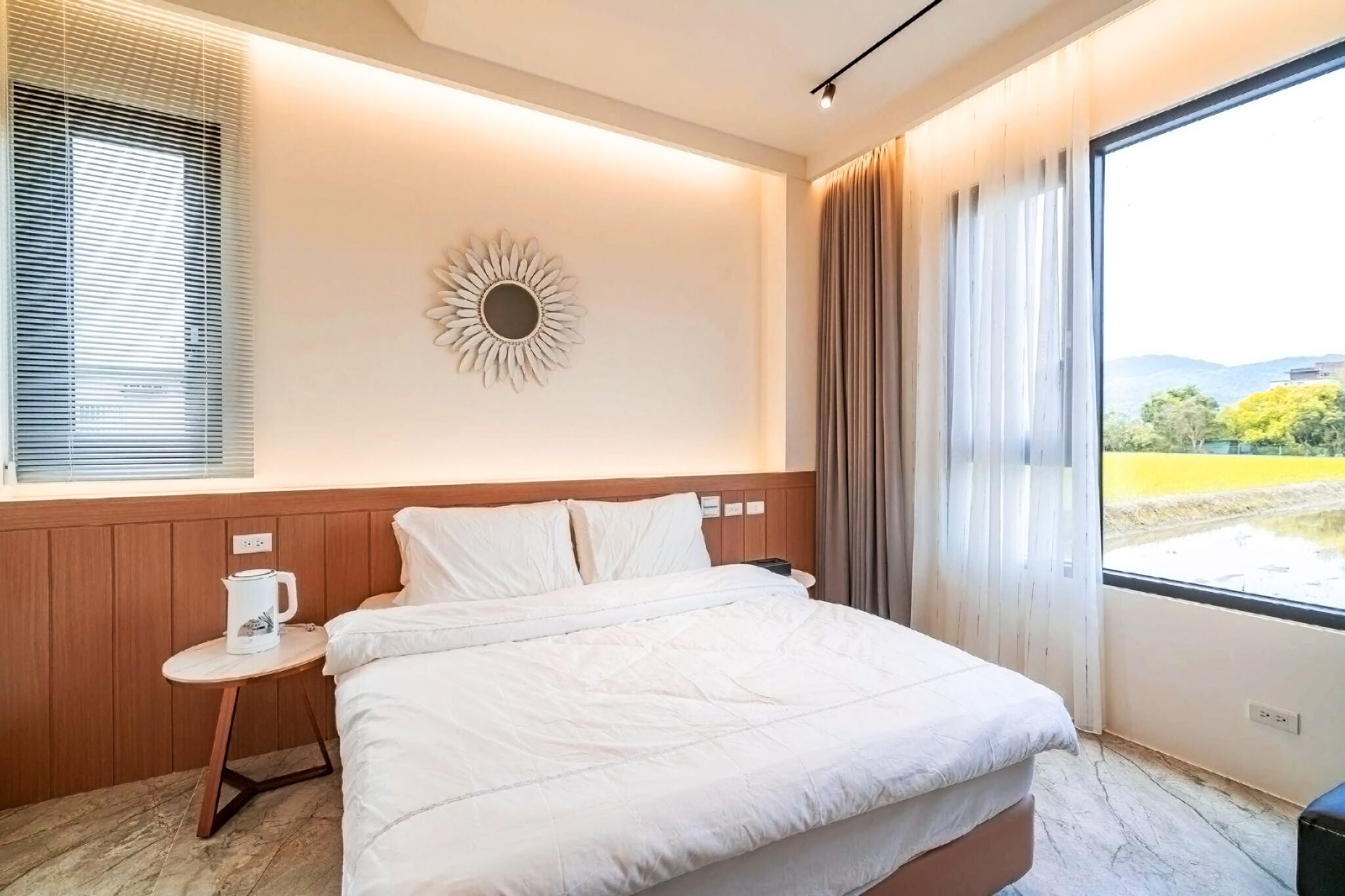 Ein Hotelzimmer mit Bett, einem kleinen Tisch mit Wasserkocher, einer Wanddekoration und einem großen Fenster mit Vorhängen.