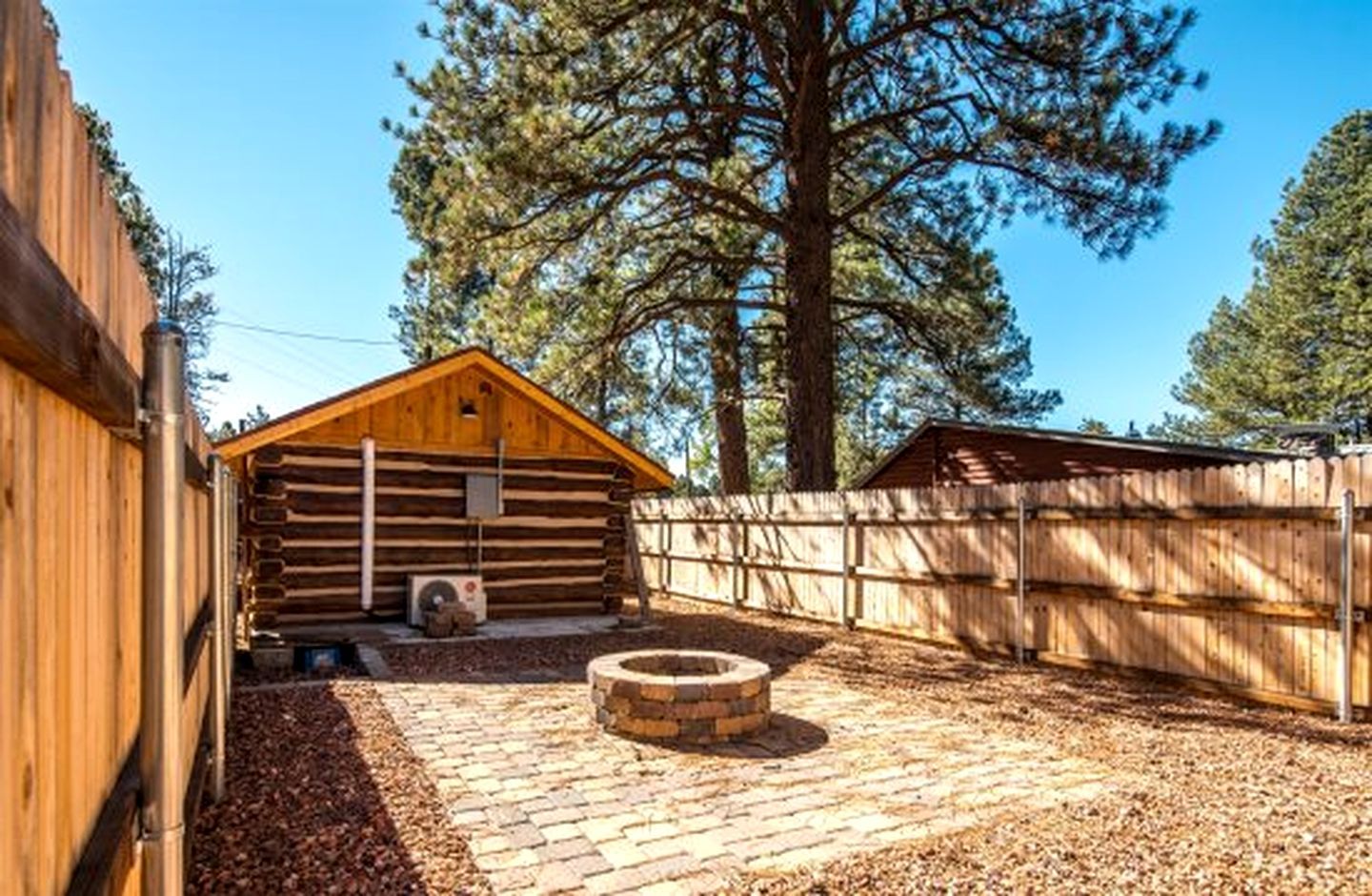 A wooden cabin with a brick fire pit in the yard, surrounded by a wooden fence and tall trees.
