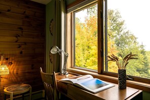 Interior de una cabaña de madera con un escritorio, una lámpara, una silla y una ventana con vistas a árboles otoñales.