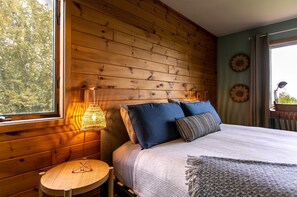 A wooden cabin-style room with a bed, a round table, and a window with a view of trees. - Family-Friendly Cabin Surrounded by Nature with Indoor Sauna in Grand Marais, Minnesota (Grand Marais)