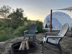 Un espace extérieur aménagé avec une tente, deux fauteuils Adirondack, un foyer et des guirlandes lumineuses.