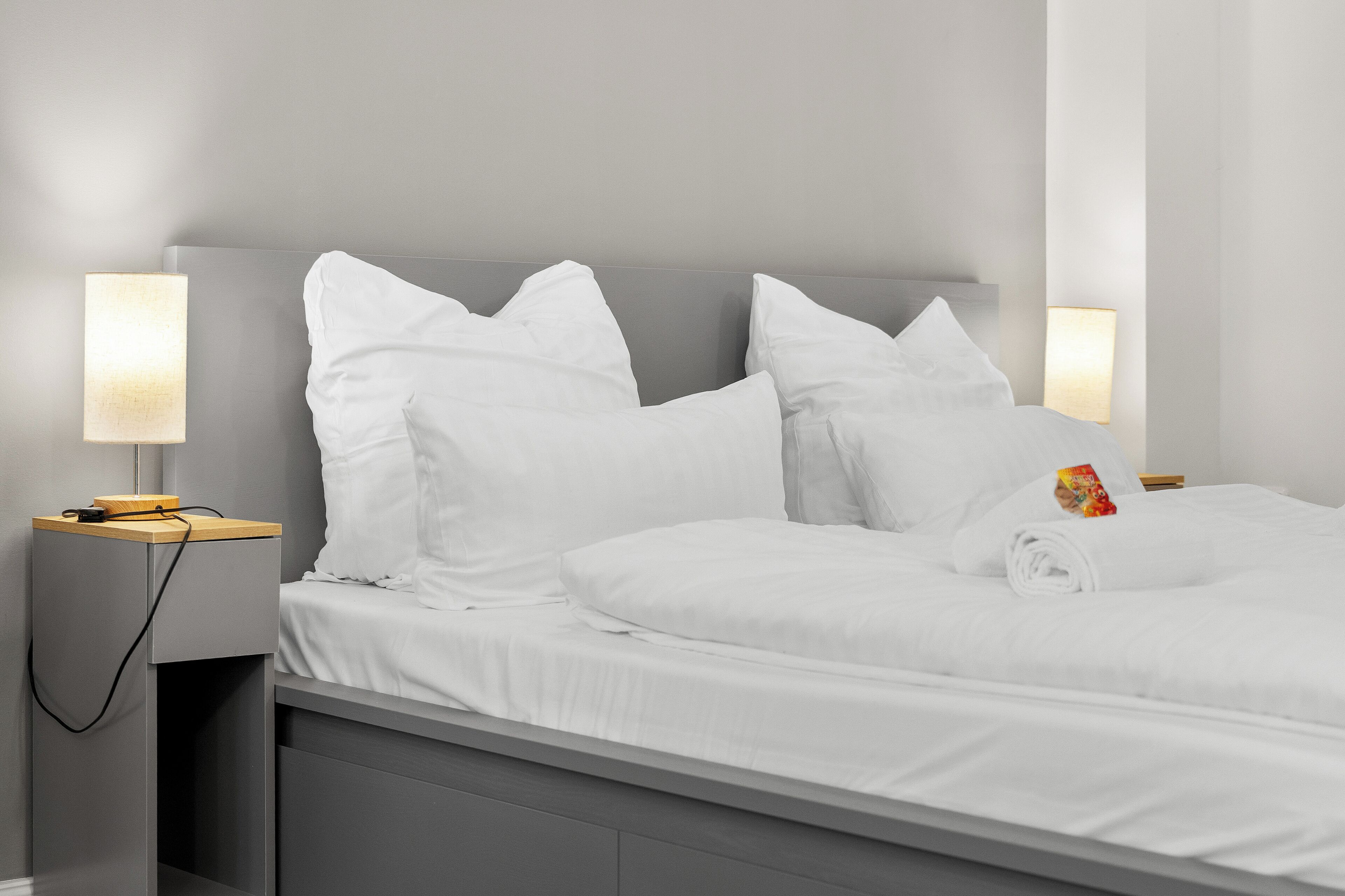 Een net opgemaakt bed met witte lakens en kussens, een houten nachtkastje met een lamp, en een handdoekje op het bed.