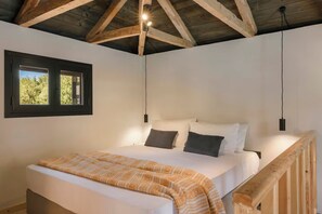 Een slaapkamer met een houten plafond, een bed met witte beddengoed en twee kussens, een raam met uitzicht op bomen, en een houten leuning.