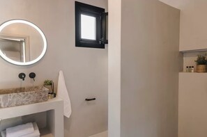 Een badkamer met een ronde spiegel, een rechthoekige spiegel, een wastafel met een marmeren blad, een handdoekhouder, een klein raam en een plank met toiletartikelen.