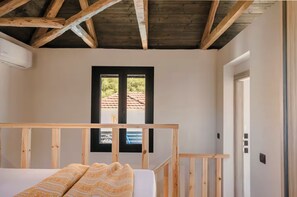 Una camera da letto con soffitto in legno, una finestra che si affaccia su un tetto con tegole rosse, un letto con coperta a motivi decorativi e una porta.
