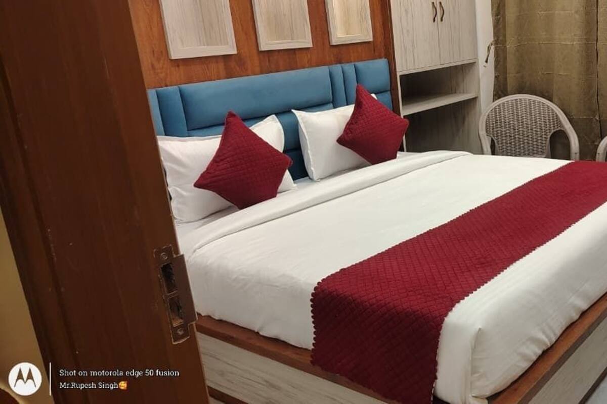A neatly made bed with a red blanket, a blue headboard, and two wicker chairs in a room with a wooden wardrobe and a curtain.