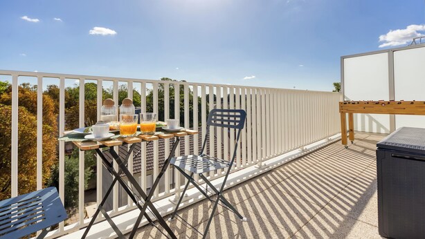Un balcon avec une table préparée pour le petit-déjeuner, une chaise et une vue sur les arbres et une clôture.