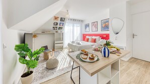 Un appartement moderne avec une chambre en mezzanine, un coin repas avec une table dressée, et un espace de vie agrémenté d’une grande plante et d’œuvres d’art accrochées aux murs.