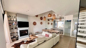A modern living room with a white sofa, a fireplace, a dining area, and a kitchen in the background. - Villa - Sleeps 8 - Near Beaches Golf and Polo (San Roque)