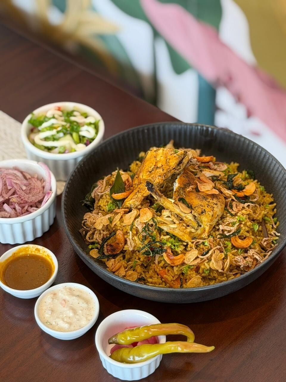 A large bowl of biryani with chicken, accompanied by small bowls of side dishes.