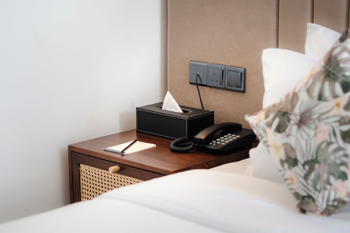 A neatly made hotel bed with a floral pillow, a bedside table with a tissue box, a notepad, and a telephone.