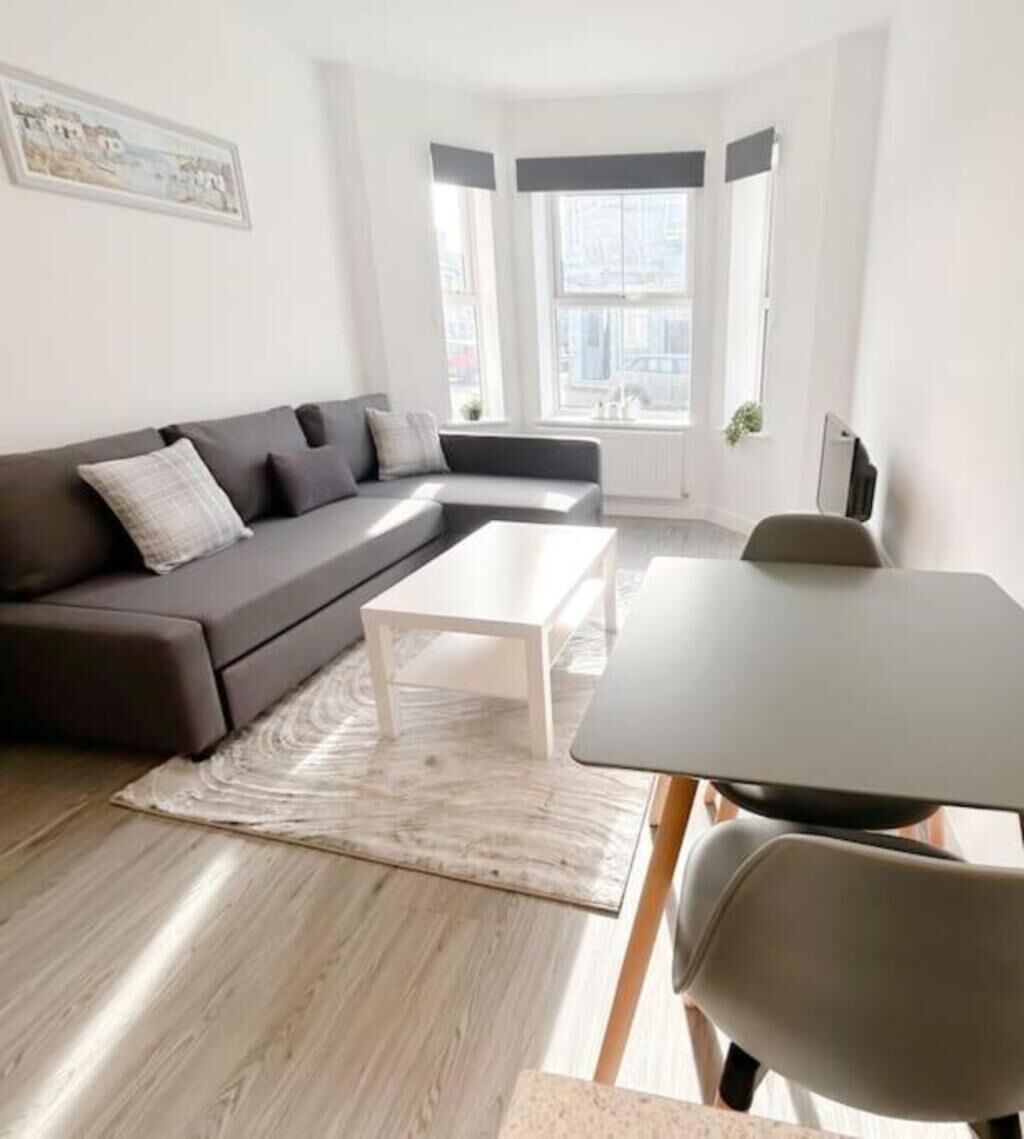 Un salon moderne avec un canapé gris, une table basse blanche et une table à manger avec des chaises. De grandes fenêtres à cadres noirs offrent une vue sur les bâtiments environnants.