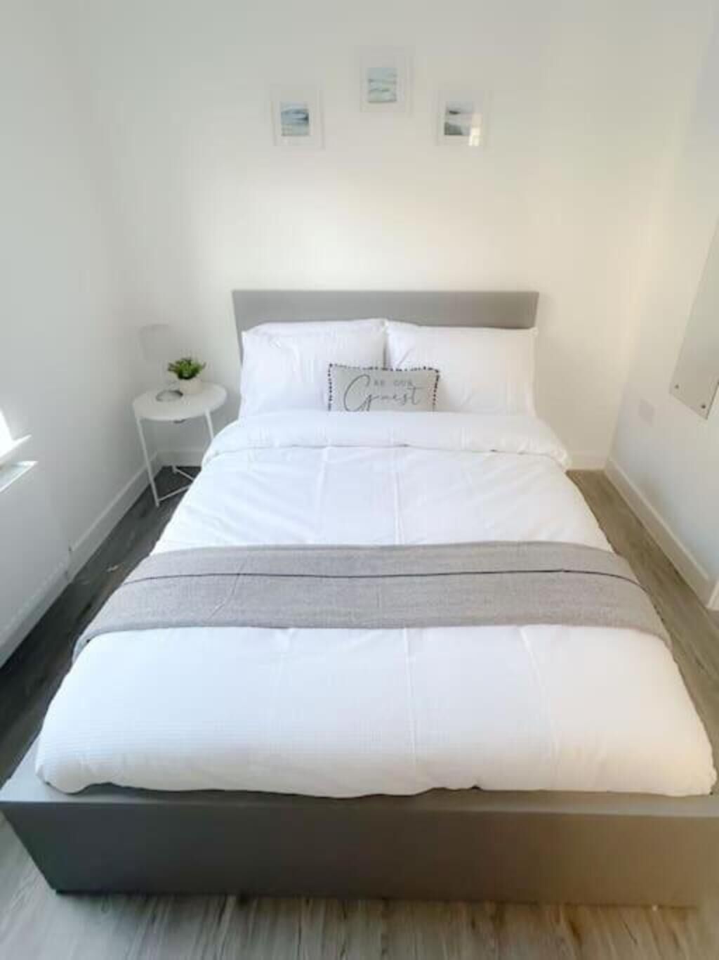 A neatly made bed with a grey headboard, a small bedside table with a plant, and framed pictures on the wall.