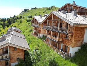 En træchalet med balkon på øverste etage, omgivet af frodig vegetation og et bakket landskab.
