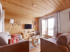 Ein gemütliches Wohnzimmer mit Holzboden, einem Flachbildfernseher, einer Holzkommode und einem großen Fenster mit Vorhängen.