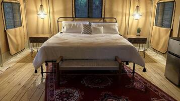 A tented room with a bed, two bedside lamps, a small table, and a refrigerator.