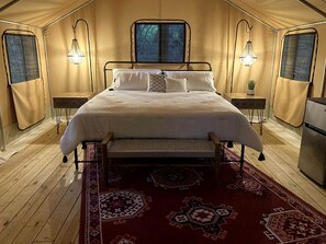 A tented room with a bed, two bedside lamps, a small table, and a refrigerator.