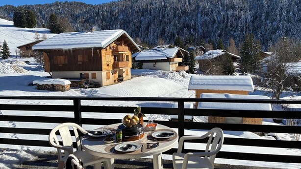 A dining table with chairs and a bowl of fruit on a balcony overlooking a snowy mountain landscape with wooden cabins. - Studio in Cernix, 4 pers, near slopes, large terrace (Crest-Voland)