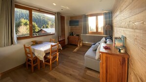 A cozy room with a wooden floor, a dining table with chairs, a sofa, and a wooden cabinet. There are windows with curtains, a radiator, and a view of trees and a house. - Studio in Cernix, 4 pers, near slopes, large terrace (Crest-Voland)