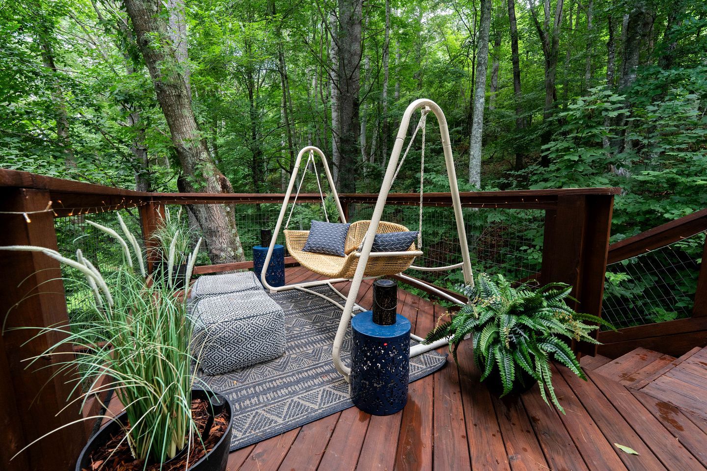 A wooden deck with a hanging chair and a small table, surrounded by greenery.