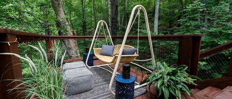 A wooden deck with a hanging chair and a small table, surrounded by greenery.