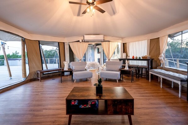 A tented room with a ceiling fan, a wooden coffee table with a floral design, two grey armchairs, a bench, and a dining table with chairs.
