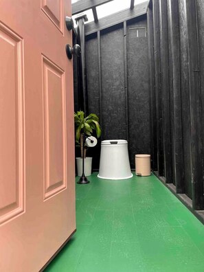 A narrow hallway with a green floor, a white trash bin, a potted plant, and a door with a black handle. - Escape to the Stars: Bell Tent Getaway for Couples in Michigan's Idyllic Destination (Boyne Falls)
