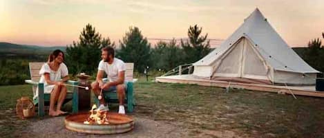 Deux personnes sont assises près d’un feu de camp dans une zone herbeuse, avec une tente en arrière-plan.