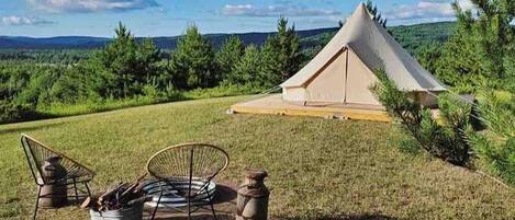 Un hébergement sous tente avec une pelouse verdoyante, deux chaises, un foyer et une vue panoramique sur les collines et les arbres.