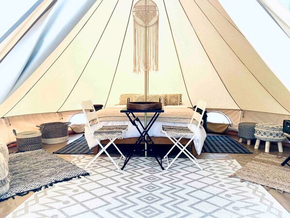 A tent interior with a central table, two chairs, a hanging light, and various rugs.