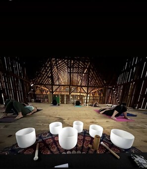 A spacious barn-like interior with a high ceiling and wooden beams, featuring yoga mats and meditation cushions arranged on the floor.