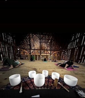 A spacious barn-like interior with a high ceiling and wooden beams, featuring yoga mats and meditation cushions arranged on the floor.