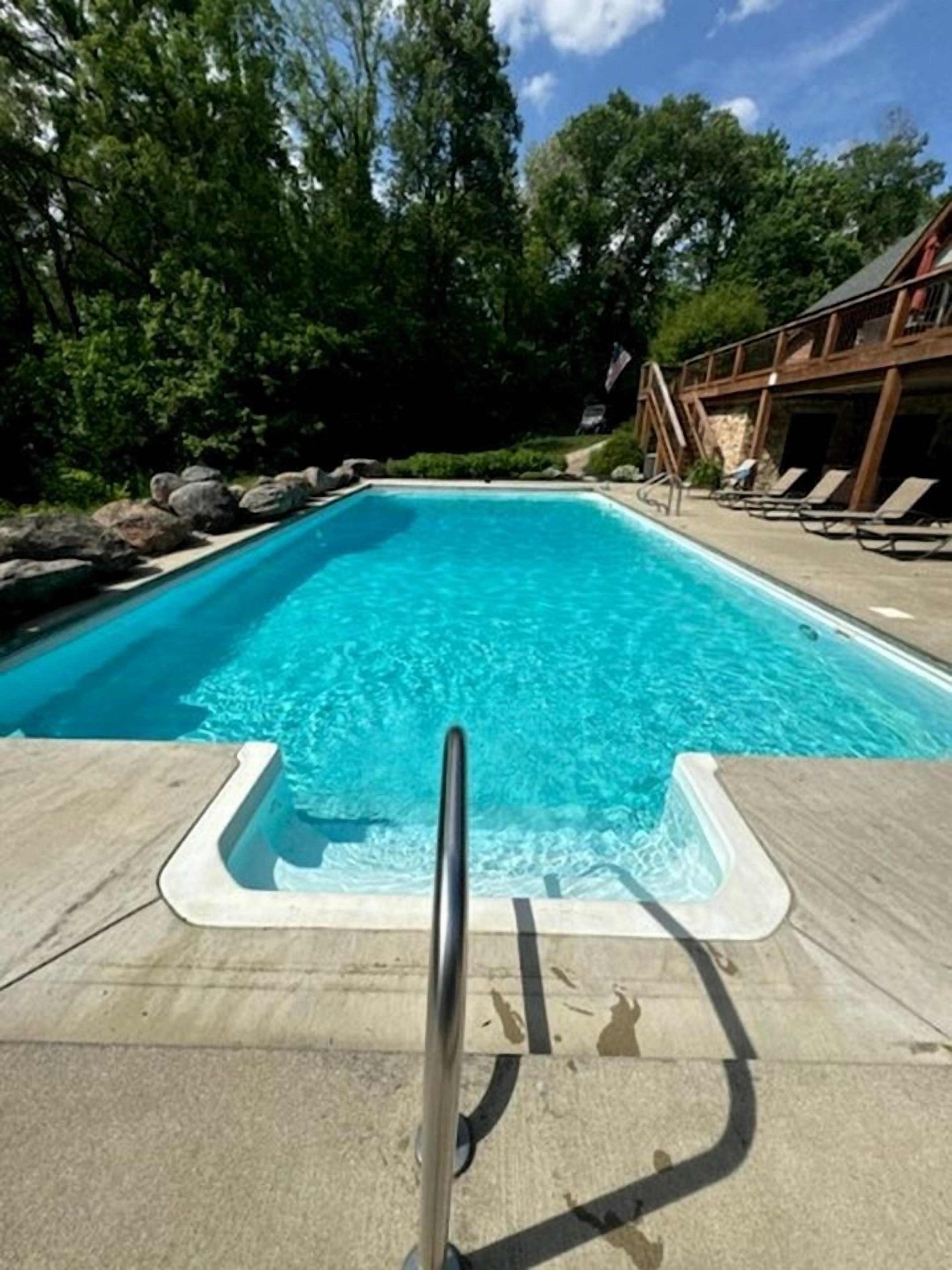 A rectangular swimming pool with clear blue water, surrounded by a concrete deck, with a wooden building in the background.