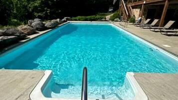 Uma piscina retangular com água azul cristalina, rodeada por um deck de concreto, e um edifício de madeira ao fundo.