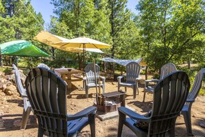 Un área de estar al aire libre con sillas Adirondack y mesas de picnic, rodeada de árboles.