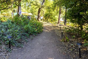 A dirt path flanked by greenery and lamp posts, leading to a shaded area with tables and chairs.