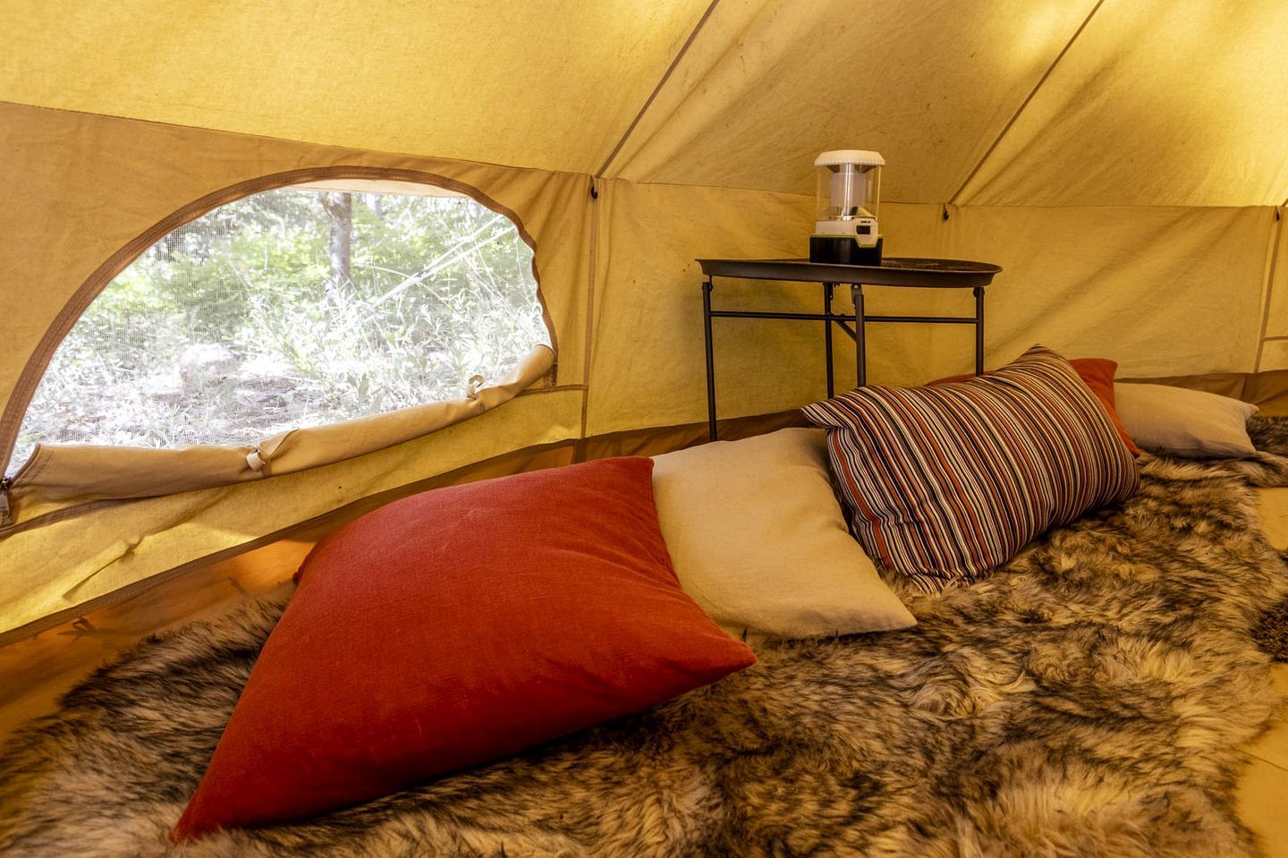 L’intérieur d’une tente, avec un lit, une table équipée d’un chauffage et une fenêtre donnant sur les arbres.