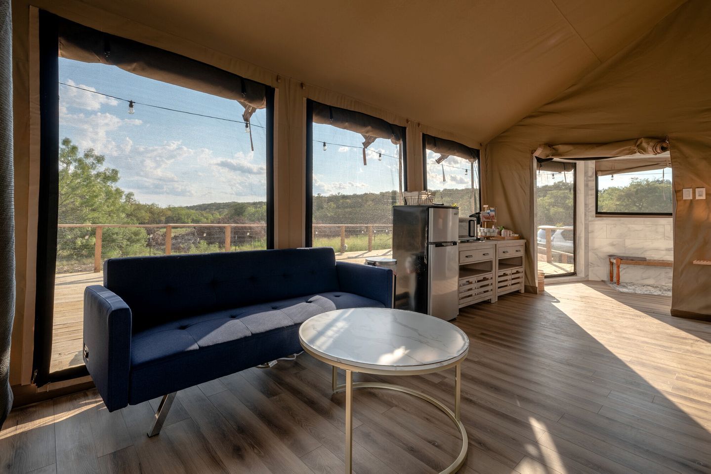 A tented accommodation with a sofa, a small round table, a refrigerator, and a view of the outdoors.