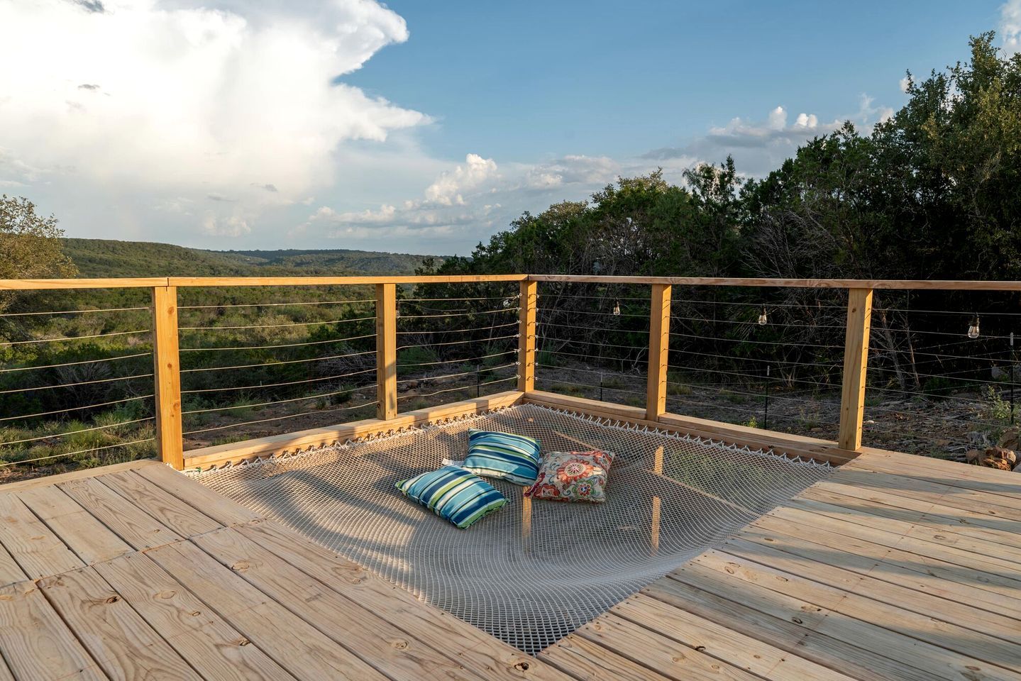 Een houten terras met een hangmat en kussens, met uitzicht op een glooiend landschap.