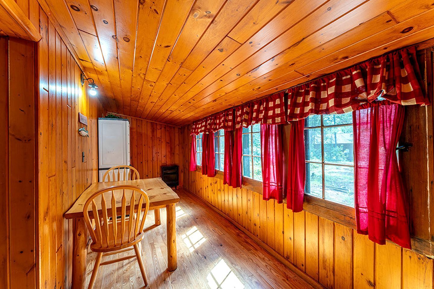 A wooden cabin interior with a dining table, chairs, and a window with red curtains.