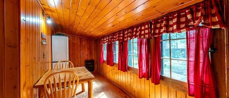 A wooden cabin interior with a dining table, chairs, and a window with red curtains.
