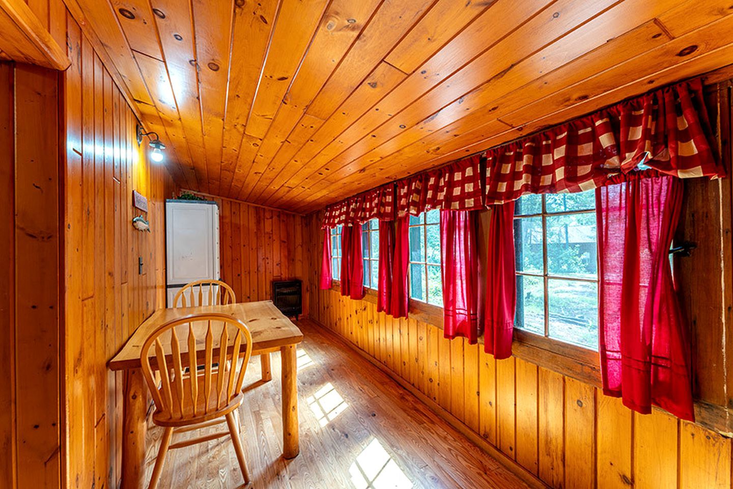 Ein Holzblockhaus-Innenraum mit Esstisch, Stühlen und einem Fenster mit roten Vorhängen.