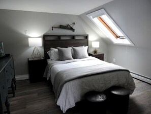 Une chambre à coucher comprenant un lit, des tables de chevet, une commode, un puits de lumière et une petite table.